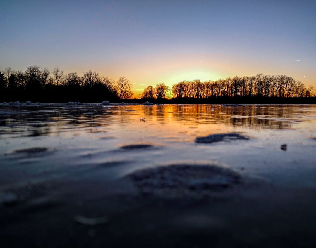 Sun sets in horizon behind a bare tree line, with lake in foreground and winter ice visible