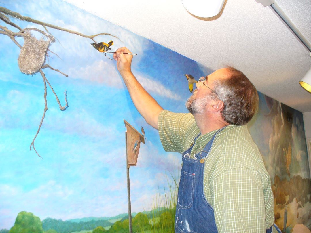 White man with gray hair, a beard, and glasses paints small details on a bird in a mural.