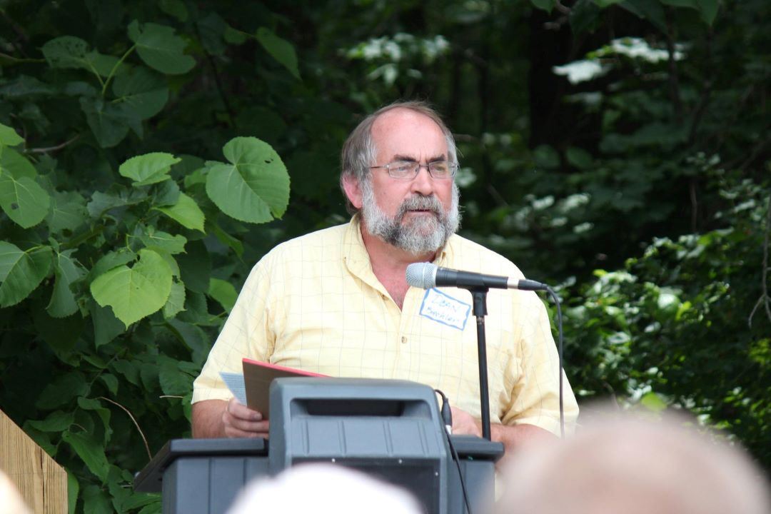 Older man with gary hair, glasses and a beard speaks into a microphone at podium