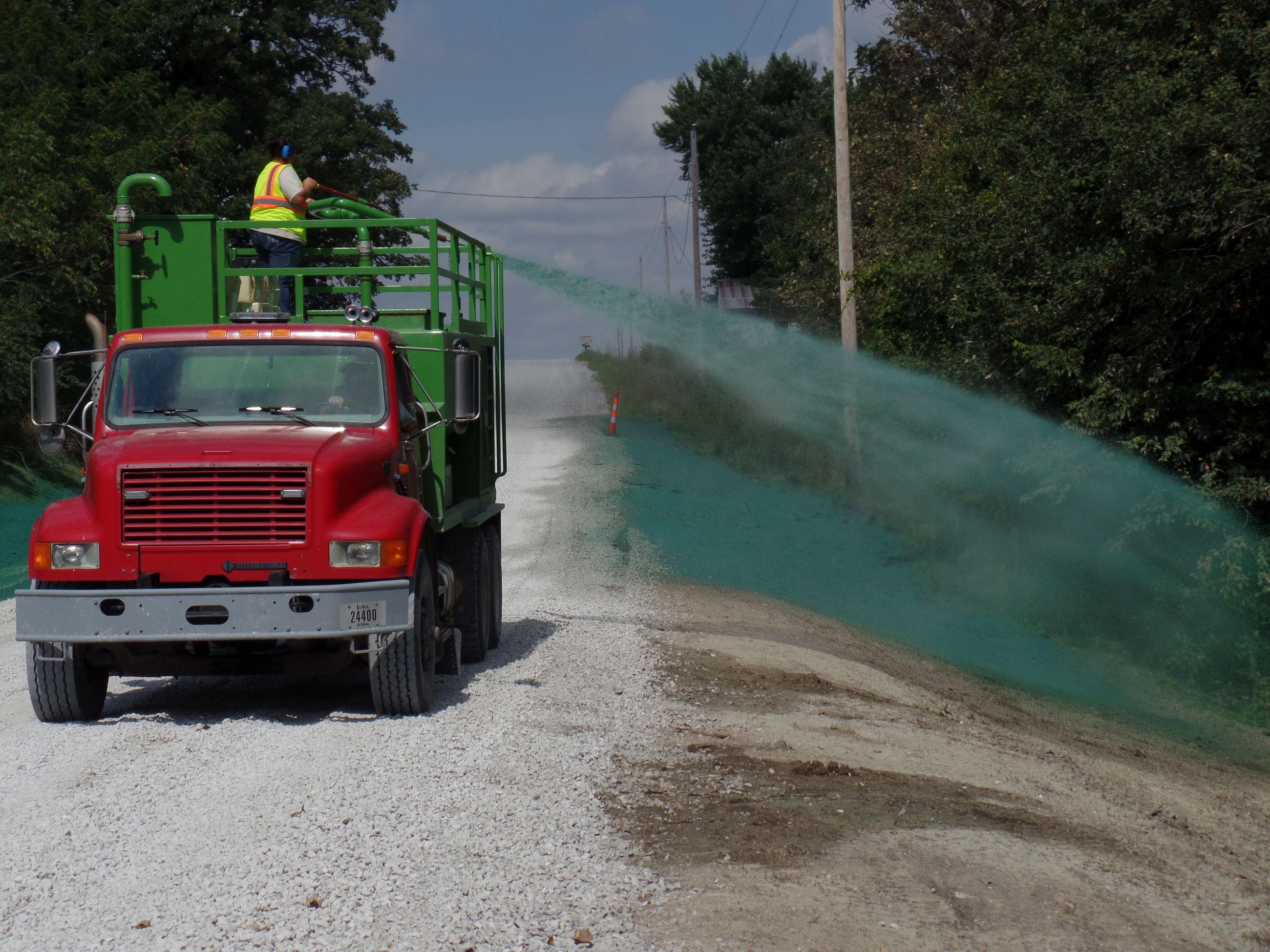 Red truck drives down gravel road, as man stands atop truck pointing green liquid spray into ditch