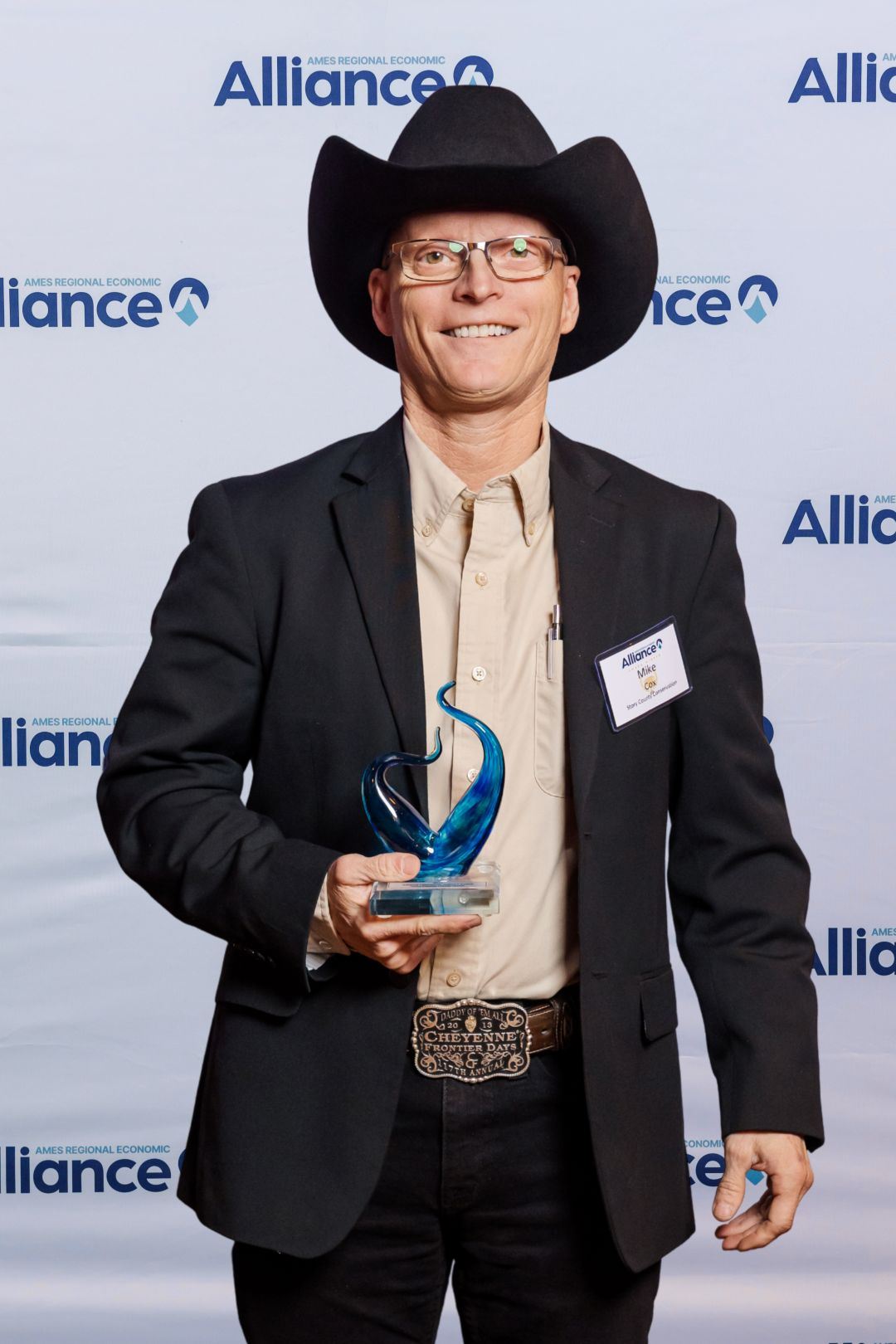Middle aged white male wearing cowboy hat holds glass award in front of photo backdrop